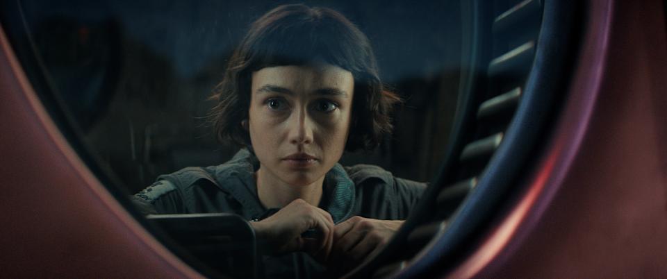A young woman, Wendy, stares pensively out a circular window in Alien: Earth, her face framed by shadows and metal, reflecting quiet resolve and underlying dread.