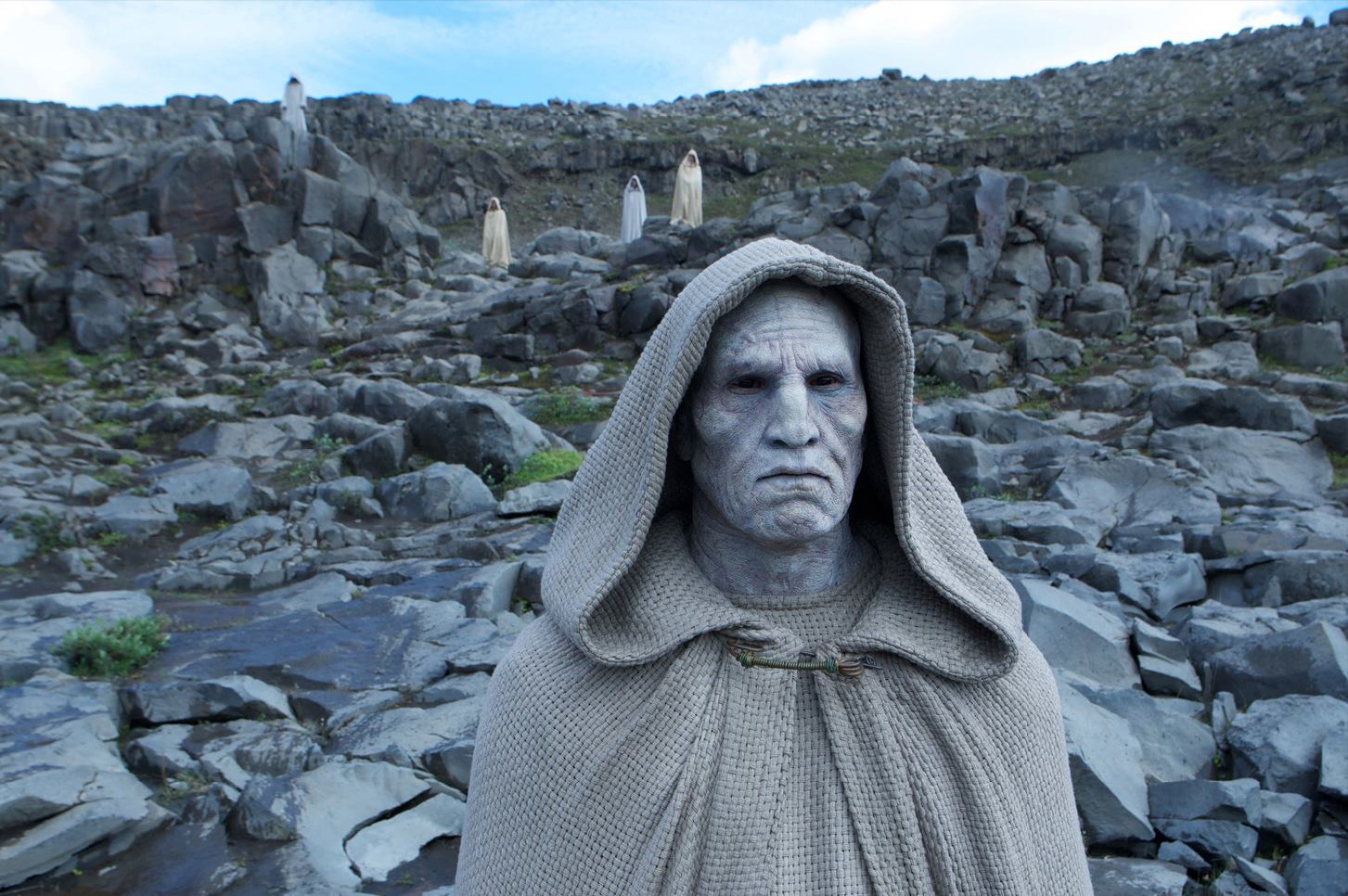A solemn Engineer cleric from a deleted scene in Prometheus, wearing a textured hooded robe and standing amid rocky terrain, with other robed figures in the background.