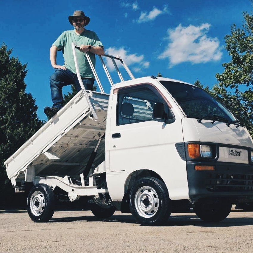 A man wearing a wide-brimmed hat, sunglasses, and a green shirt stands confidently on the tilted dump bed of a small white kei truck under a bright blue sky. The truck is parked on a sunny day with trees in the background.