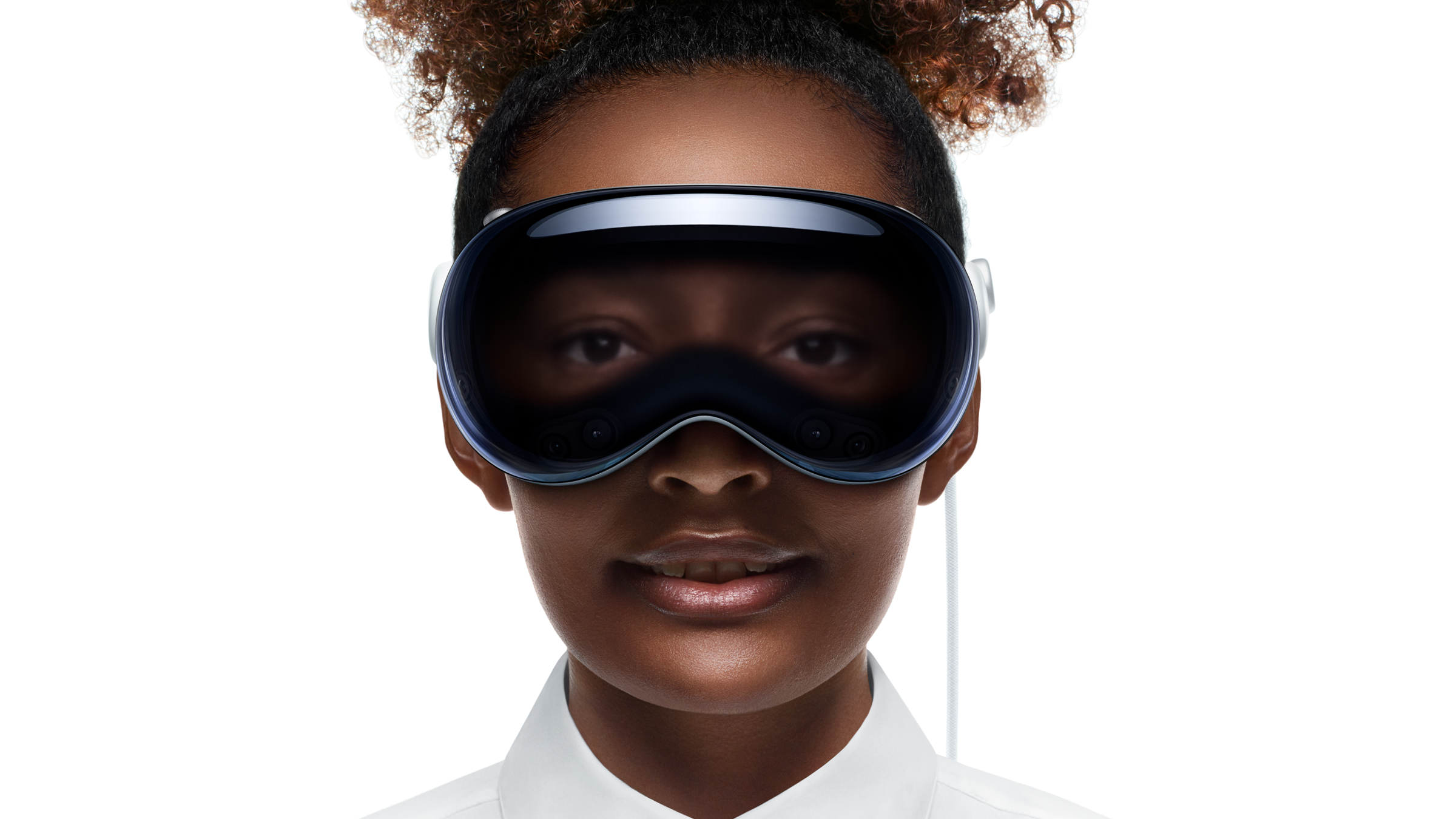 This is a promotional photo from Apple showcasing a young woman wearing the Apple Vision Pro, their mixed-reality headset. The woman is depicted from the shoulders up, with her face centered in the frame. She has dark skin, a natural hairstyle with curls tied up, and is wearing a crisp white shirt. The Apple Vision Pro covers her eyes and upper face. Its sleek design features a dark, reflective, curved visor and a soft inner lining. The headset's front surface displays an augmented projection of the wearer’s eyes, creating a digital effect where her gaze is visible through the visor. The image highlights the futuristic and human-focused aspects of the technology.