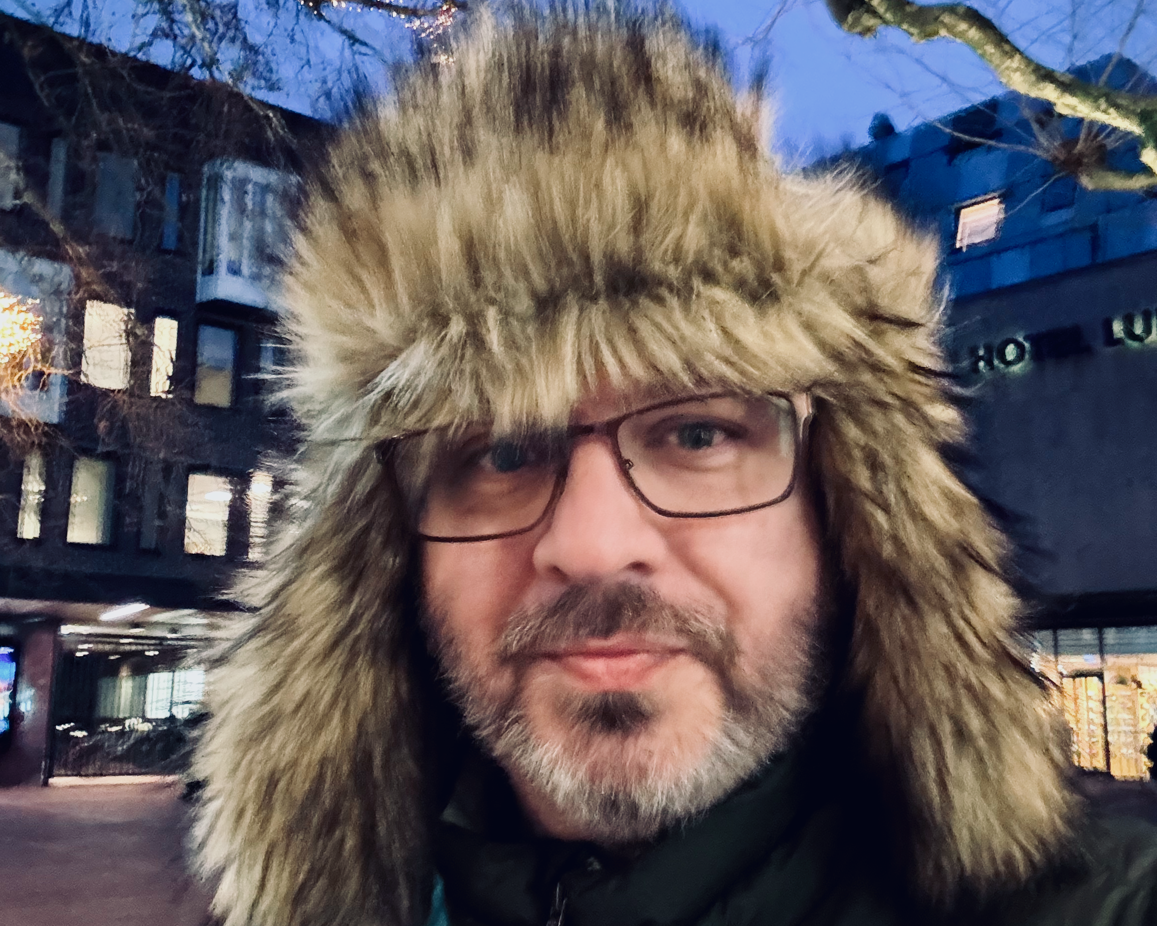 Selfie of Magnus, a middle aged white man with glasses and a beard. He is wearing a big furry arctic hat with ear flaps. It's evening outside, and there are bits of a blurry urban Swedish scene in the background.